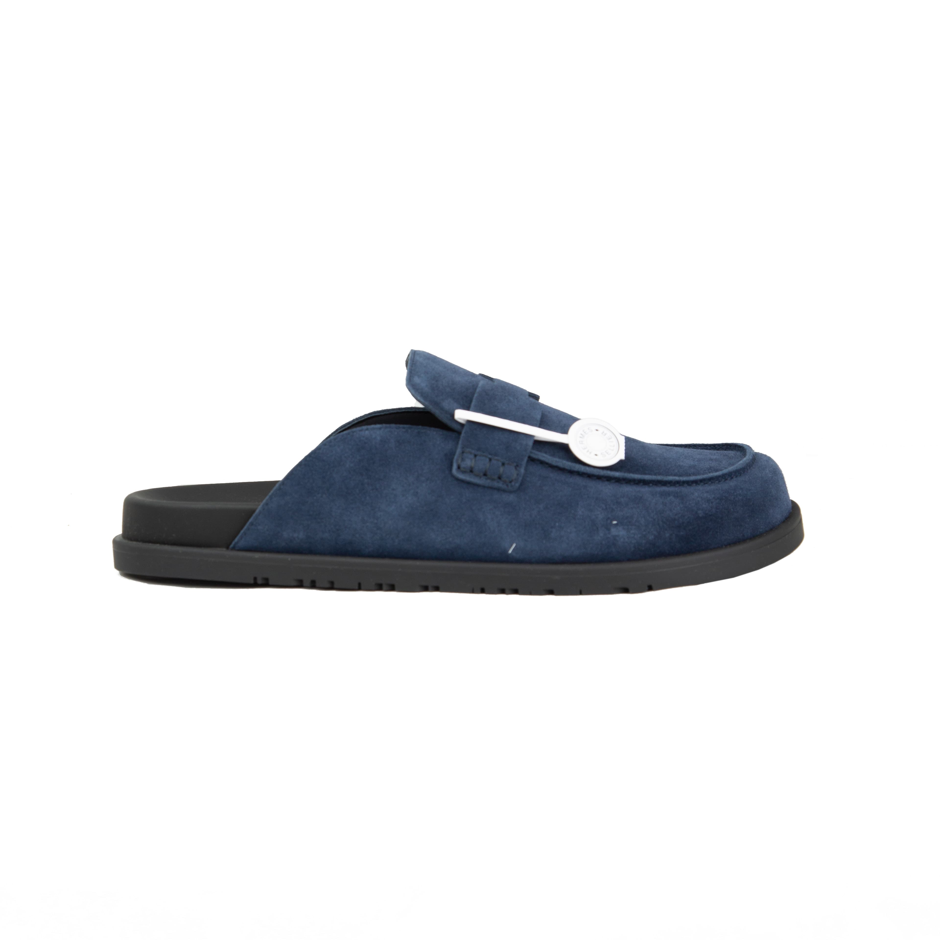 Pair of navy blue suede Hermès Ancora mules with tonal H strap and black rubber soles on a white background.