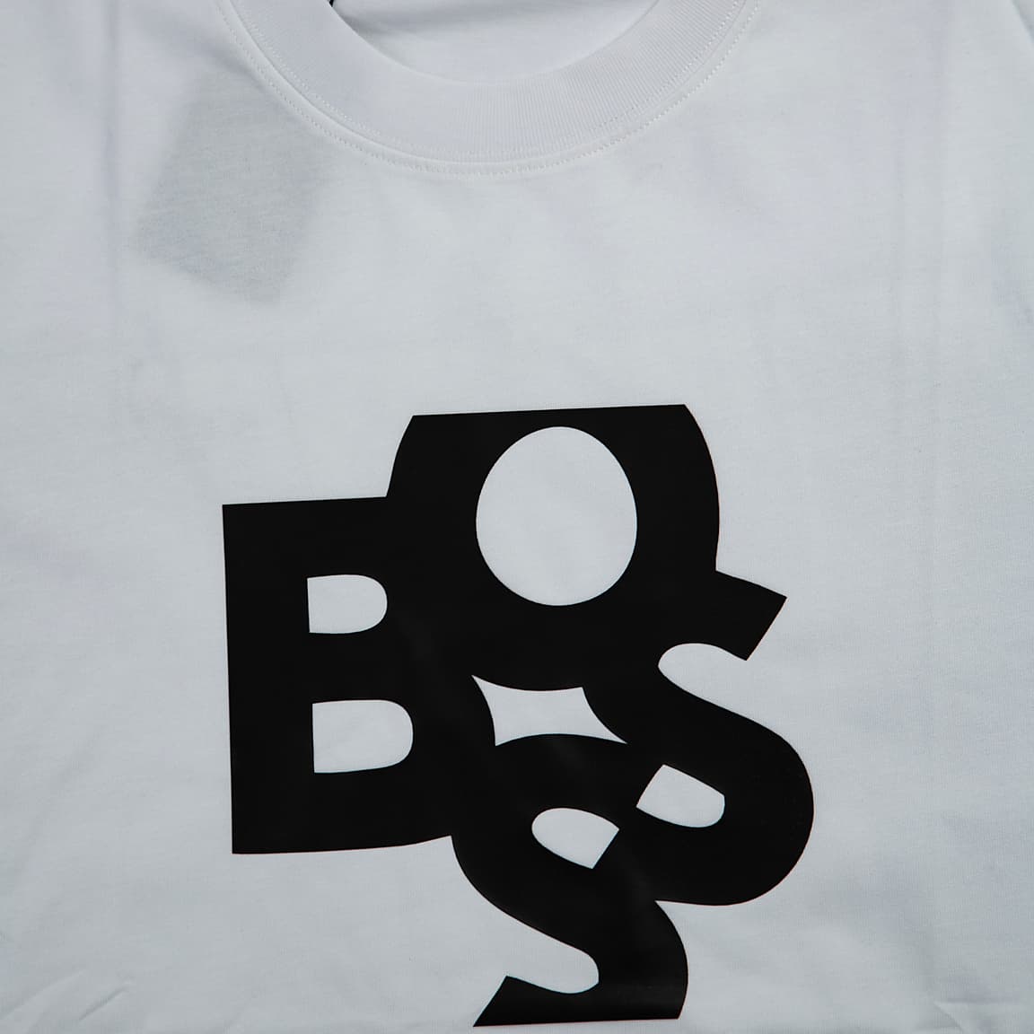 A simple, modern light grey (or very pale off-white) short-sleeved, crew-neck t-shirt. It features a bold, large black graphic of the word "BOSS" rendered in a stylized, abstract, and overlapping script across the chest.