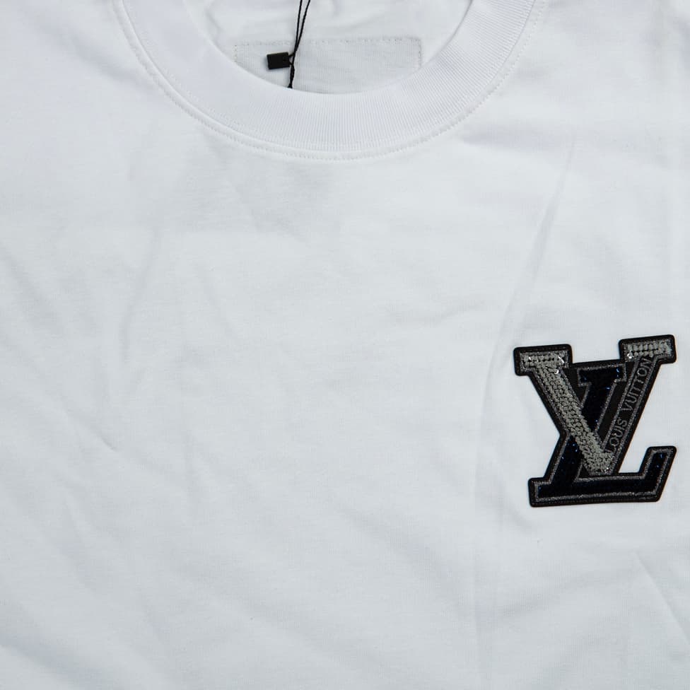 A clean, plain white, short-sleeved, crew-neck t-shirt. It features a small, striking appliqué patch on the left chest.