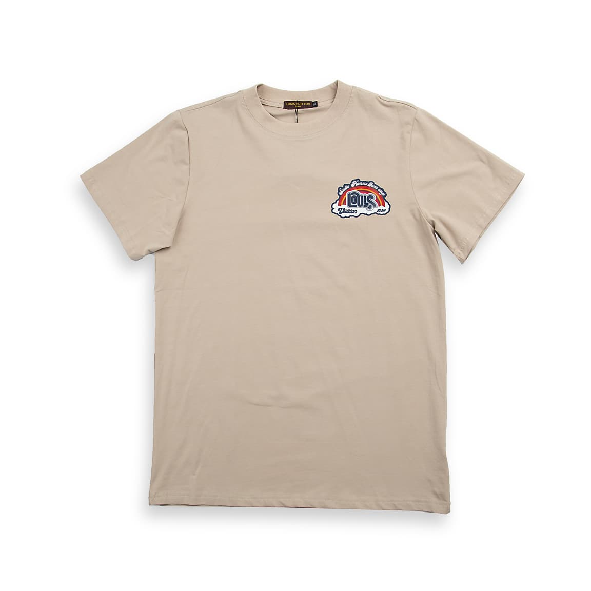 A light, neutral beige (or taupe/tan), short-sleeved, crew-neck t-shirt. It features a small, colorful, embroidered or printed graphic patch on the left chest.