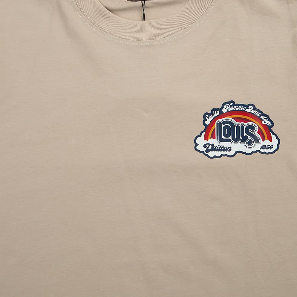 A light, neutral beige (or taupe/tan), short-sleeved, crew-neck t-shirt. It features a small, colorful, embroidered or printed graphic patch on the left chest.