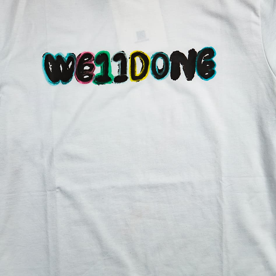 White WE11DONE graphic t-shirt with glitch logo, short sleeves, streetwear style, laid flat.