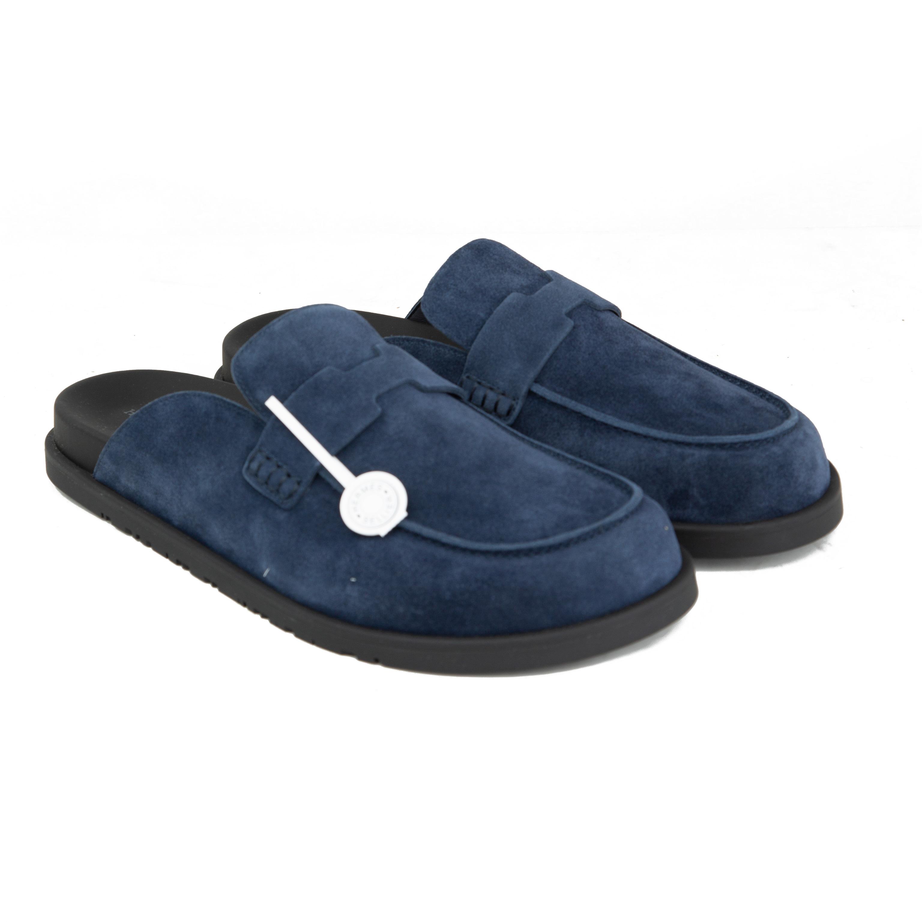Pair of navy blue suede Hermès Ancora mules with tonal H strap and black rubber soles on a white background.